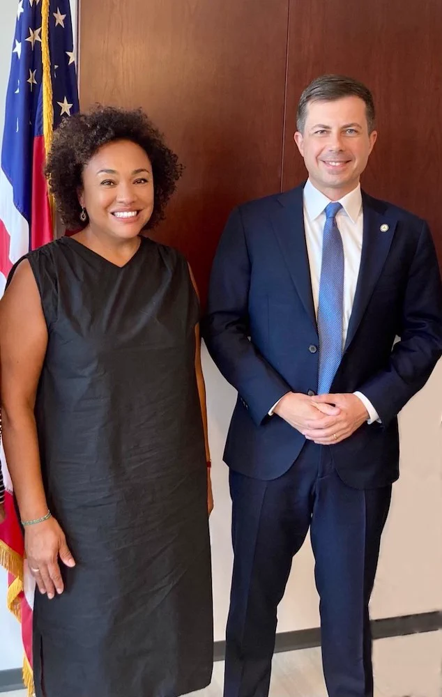 Maria Rosario Jackson, who is an Afro Latina woman, with Pete Buttigieg, who is a white man. Maria is wearing a mid-length black linen shift. Pete is wearing a blue suit with a lighter blue tie. They are both smiling at the camera.