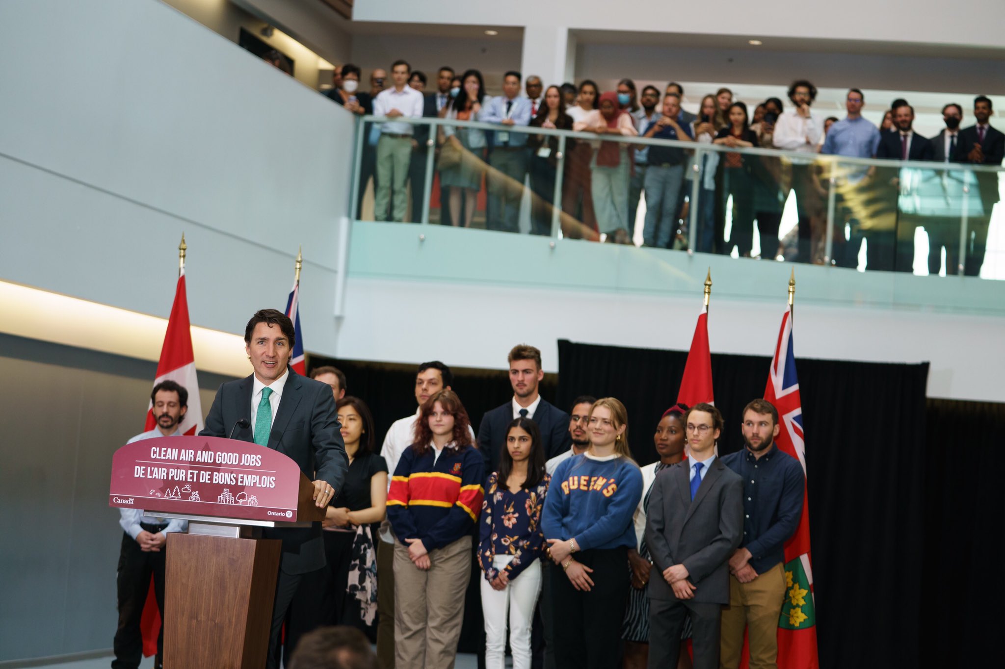 Prime Minister Justin Trudeau is standing at a podium and speaking. A large group of students are standing behind him. In the background, more students are standing on the second level of the atrium.
