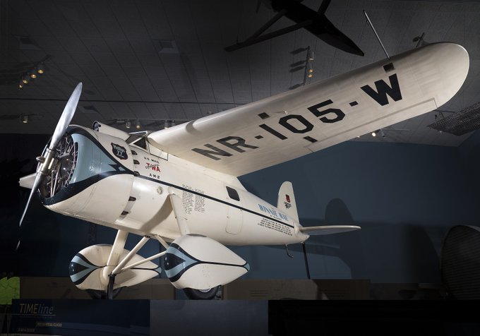 Lockheed Vega Winnie Mae, a white and blue aircraft, hanging in a gallery. 