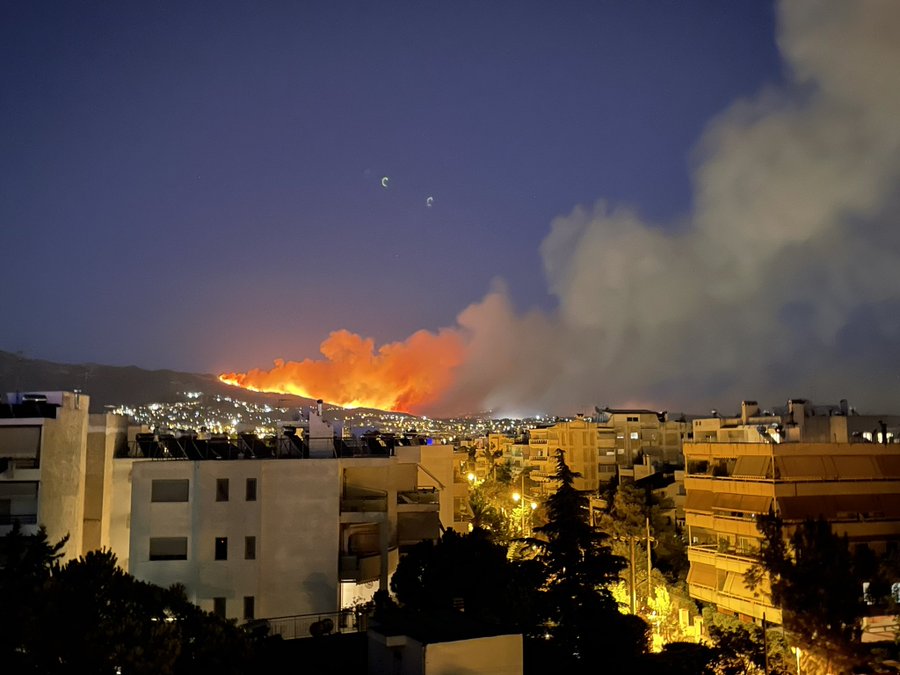 A wildfire rages in the back part of photo with huge clouds of billowing  smoke. it’s twilight, and urban housing blocks typical in Athens  Greece fill the rest of the photo.