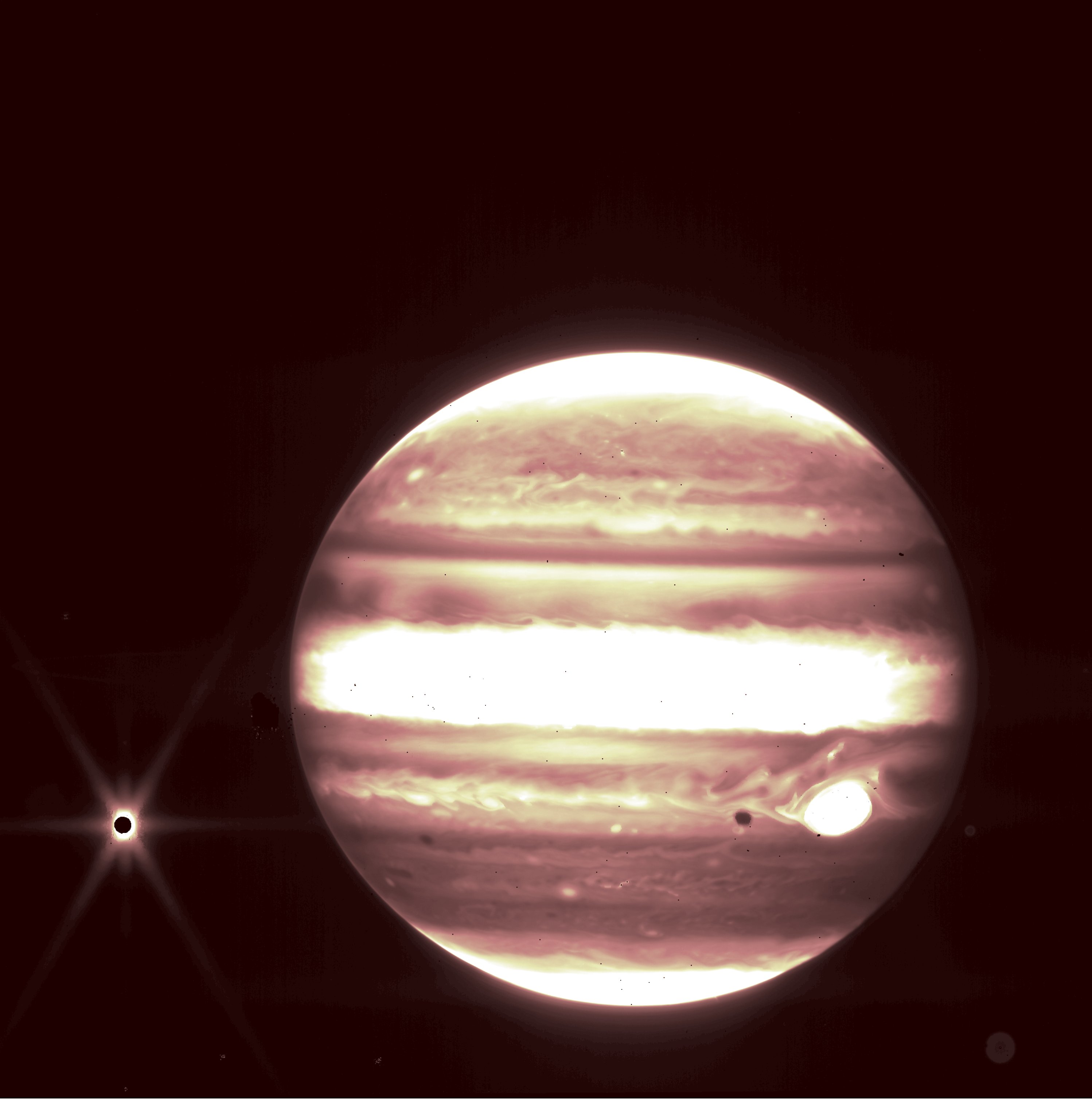 Jupiter dominates the frame, appearing to glow with bands of bright white, light yellow, and darker, brownish oranges. The stripes circle the planet, with one especially thick bright band across the planet’s center. A spot of glowing bright white interrupts the darker brown band about a third from the bottom of the planet. To the left of Jupiter, Europa appears as a tiny, black circle with a bright starburst erupting from its edges. The background of the image is pure black.