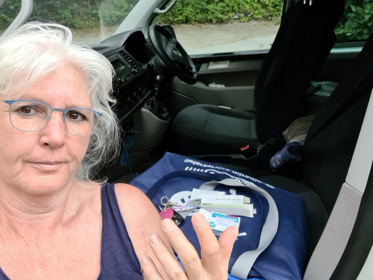 Frau steht am offenen Auto, im Hintergrund Grün.<br>Innen auf dem Sitz Antibiotika-Schachteln, sie hält die Hand hoch, der rechte Zeigefinger ist ziemlich dick.