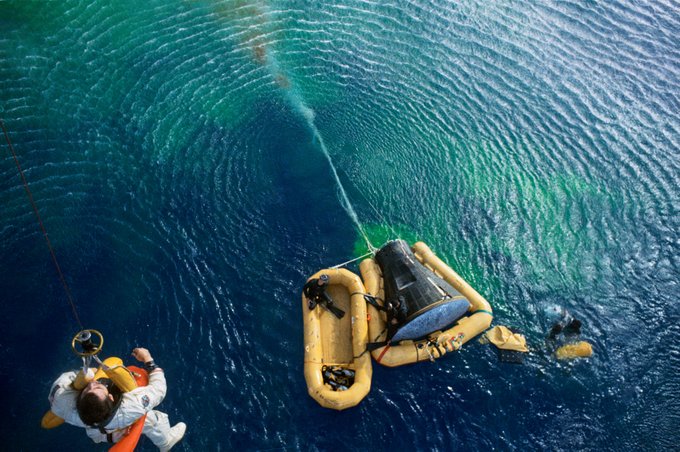 On a backdrop of blue-green rippling Atlantic Ocean water, astronaut John Young is visible in the bottom left corner being raised up on a cable to the recovery helicopter. In the bottom middle, the Gemini X spacecraft is above the surface of the water, floating on top of yellow flotation devices. In one floating raft, a person in black scuba gear is looking up as Young rises in the air.