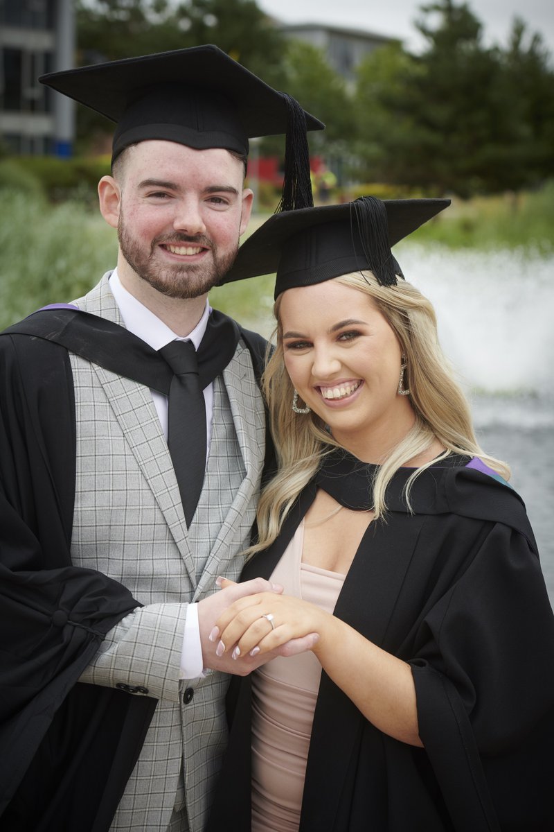 Two people in graduation robes smile for the camera, one showing an engagement ring on their finger