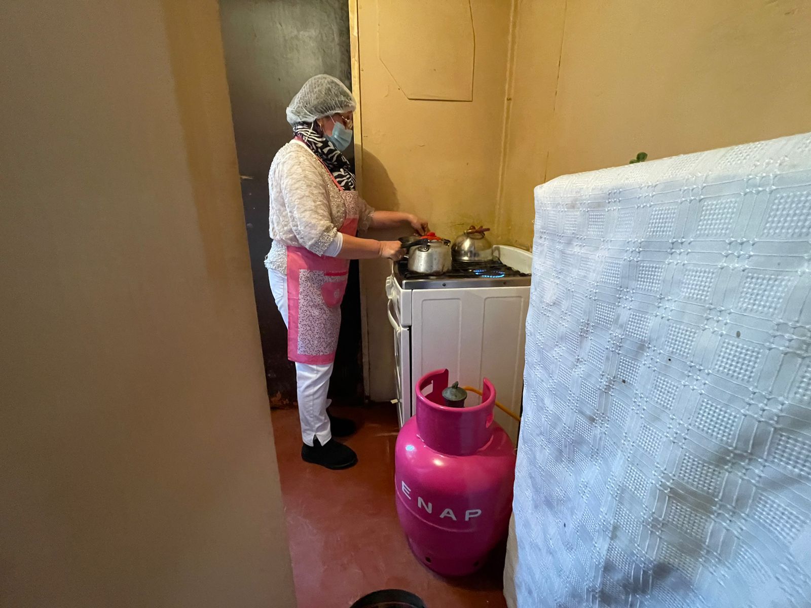 Mujer con malla en la cabeza y delantal, hirviendo agua con su cilindro rosado de Gas de Chile.