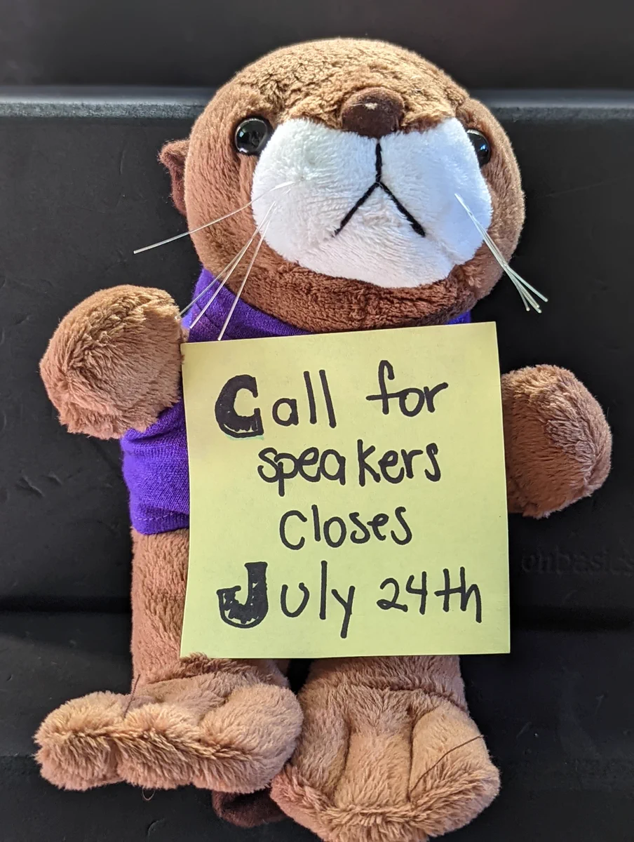 A stuffed toy of Mani the otter, holding a sing that says "Call for speakers closes July 24th"