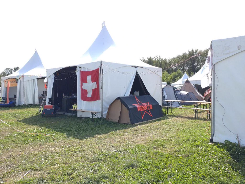 The tents of the MCH2022 villages (from left to right) UAVP, SwissVillage and Lemanicus with a Swiss flag hanging outside the SwissVillage tent and a Keyboard Star flag hanging over a KarTent.