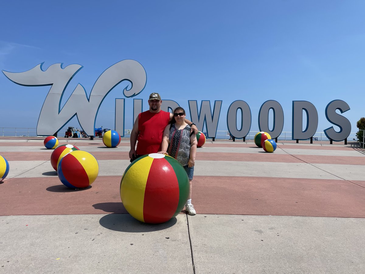 Just a couple of lovebirds on vacation...🏖️

[Image description: Amanda and Greg are shown arm in arm, smiling for the camera in front of a Wildwoods banner overlooking the ocean].
