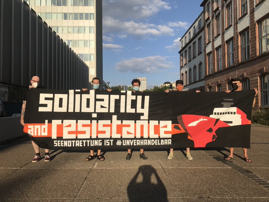 5 Menschen halten ein großes schwarzes Transpi<br>auf den linken 2/3 steht <br>[Lettern in weiß]: solidarity <br>and resistance [auf einem orangen Streifen] <br>SEENOTRETTUNG IST <a class=