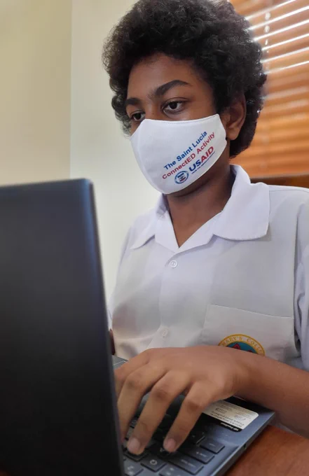 Student types on laptop computer wearing a white shirt and white mask that says "The Saint Lucia ConnectEd Activity" with the US Agency for International Development (USAID) logo.