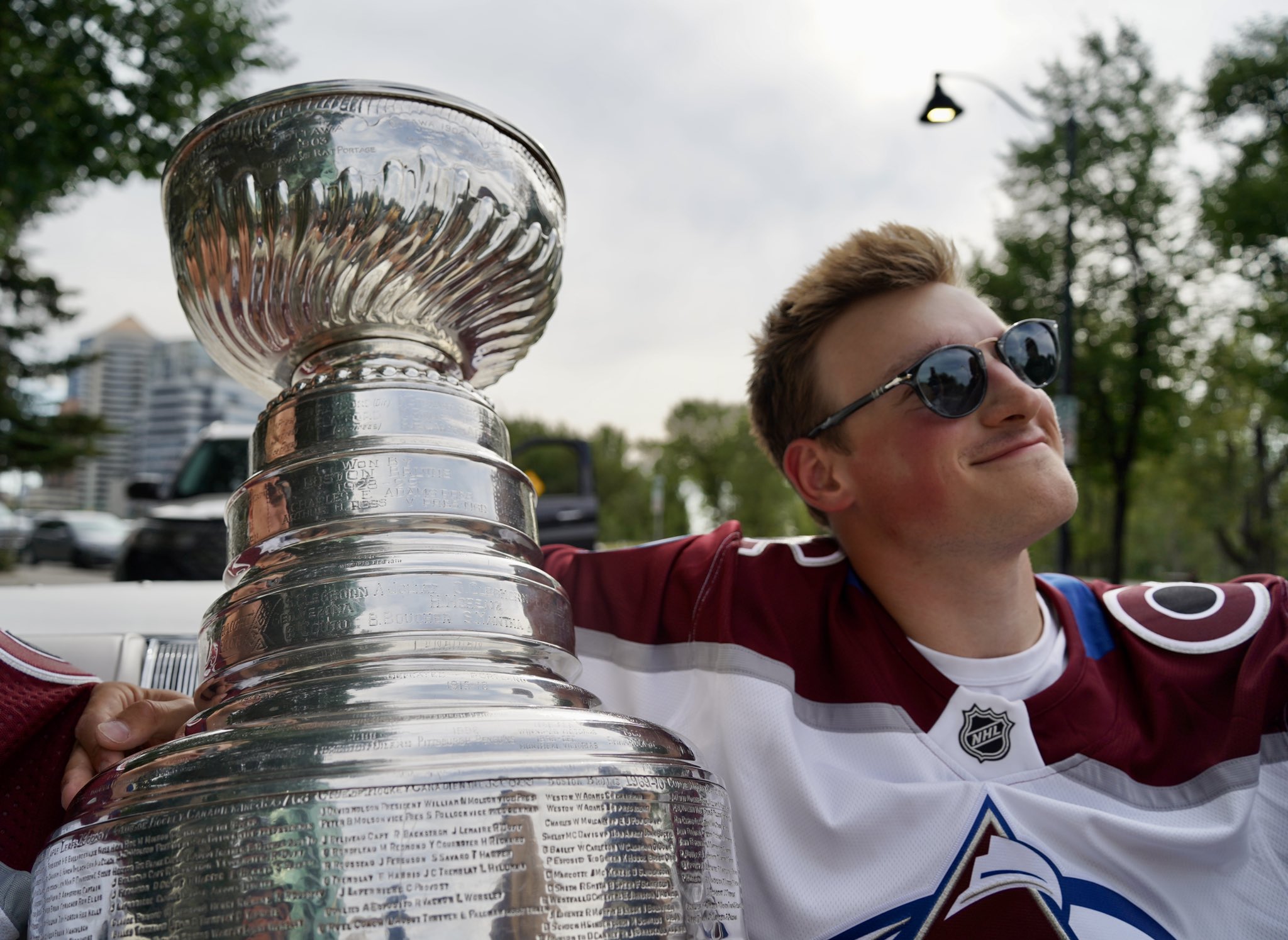 Cale Makar soft smiling with his arm around the Stanley Cup in a convertible. He is wearing his Avalanche jersey and has sunglasses on.