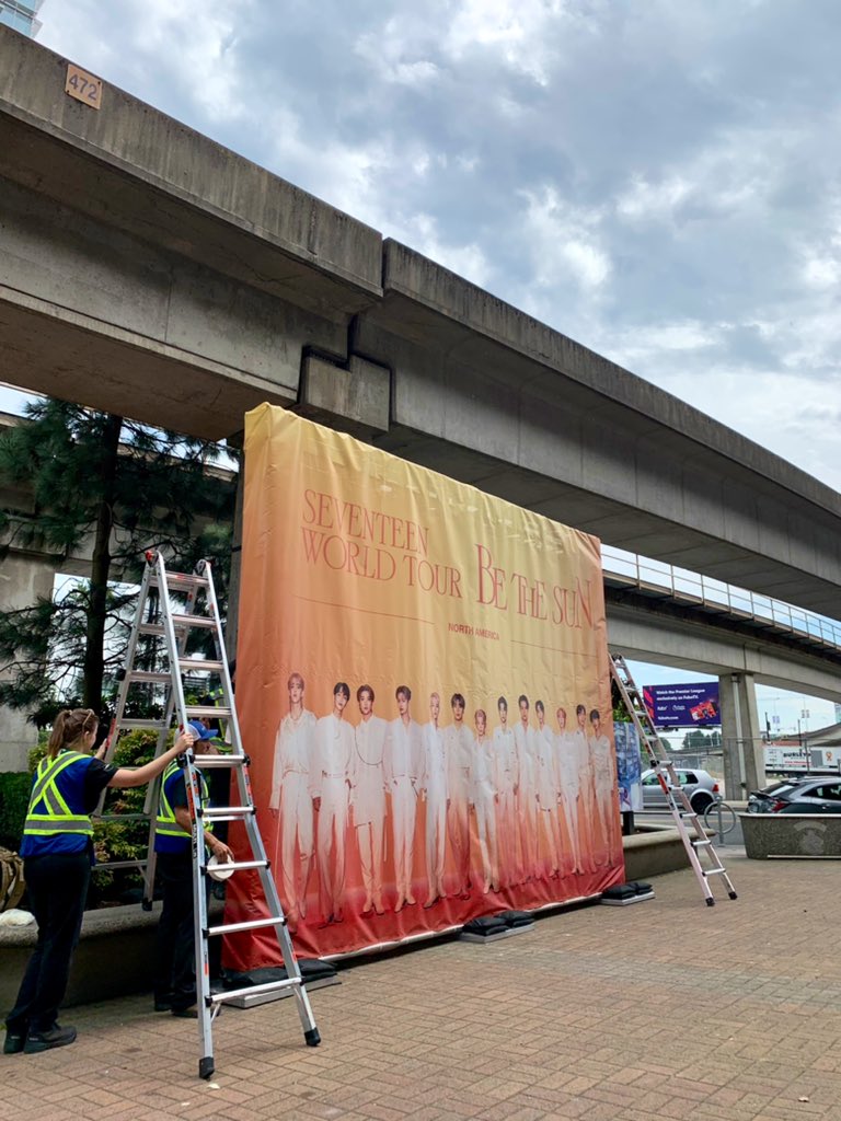 Photo of personnel setting up a Seventeen World Tour: Be The Sun standee at the North Plaza in front of Rogers Arena - it portrays each member wearing white outfits, with the name of the tour written above them.