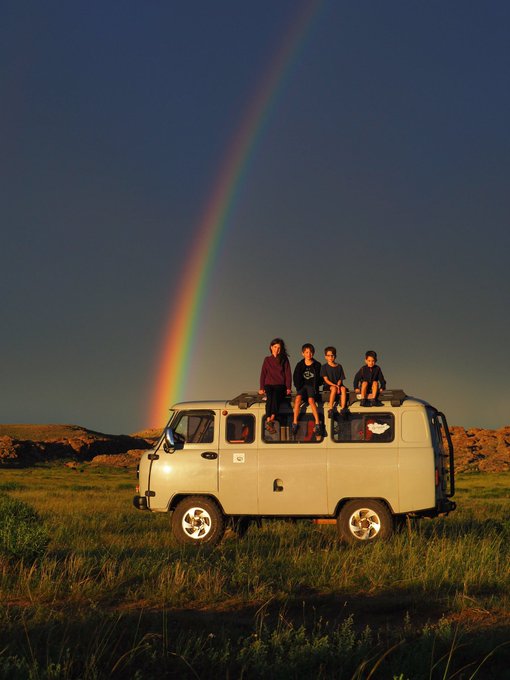 4 enfants assis sur le toit d'une camionnette grise.  Ils sont éclairés par la lumière orangée du soleil couchant. Derrière eux on voit un magnifique arc-en-ciel.