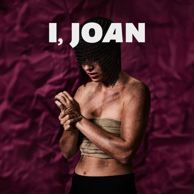 A graphic of a person who is covered in dirt and grime, their chest is bound and they wring their hands. A chainmail helmet covers their eyes. They stand in front of a purple background with text above their head reading I, JOAN in large white letters.