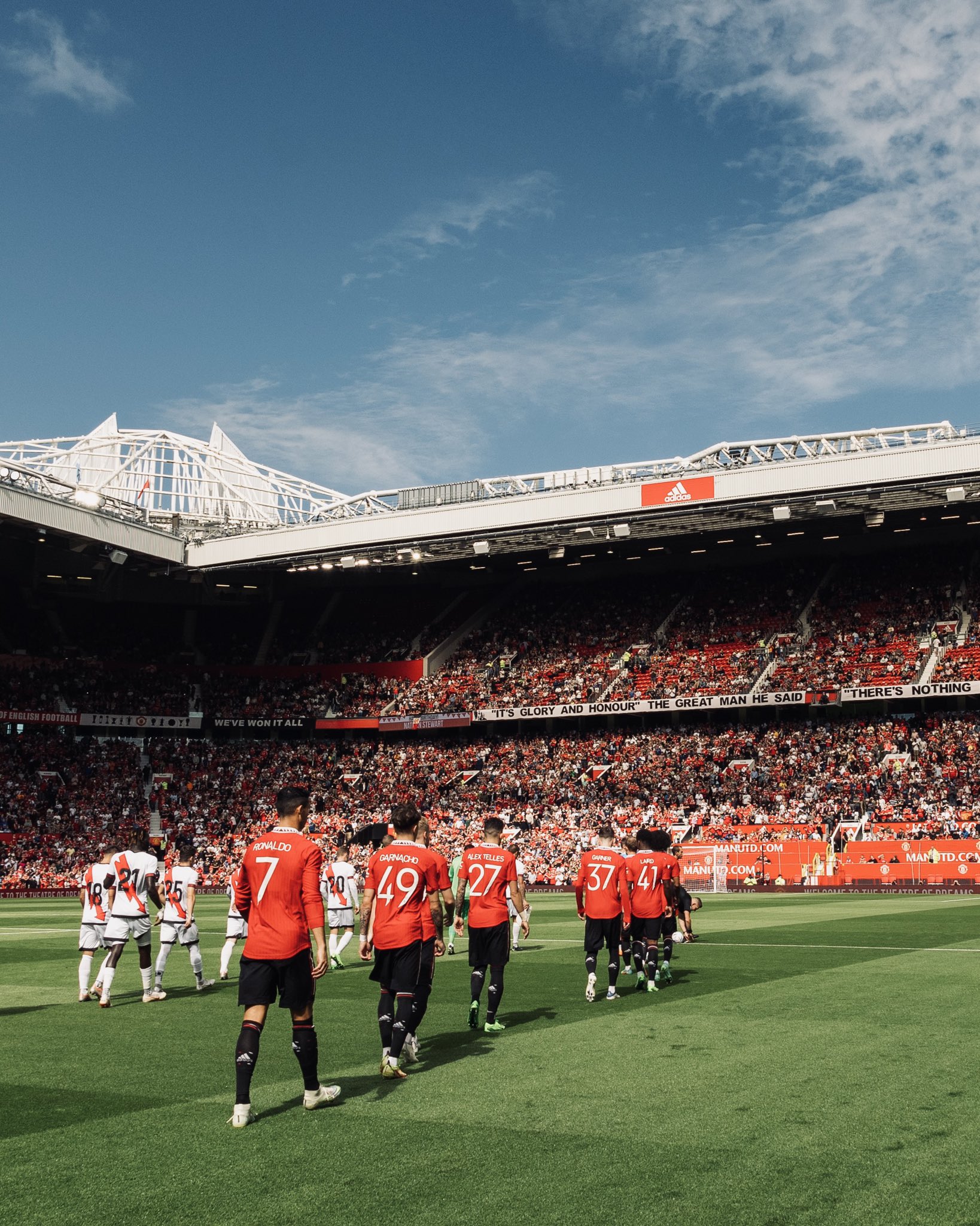United players walkout onto the pitch alongside Rayo Vallecano, with the East Stand in the background beneath a blue sky.