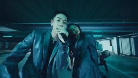 Two performers in a dimly lit parking garage sing into a microphone during a music video shoot with blue-tinted lighting.