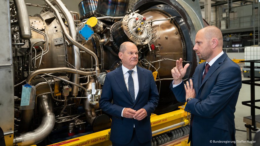 Bundeskanzler Olaf Scholz steht mit dem Vorstandsvorsitzenden von Siemens Energy Christian Bruch in einer Fabrikhalle vor einer großen Gasturbine. 