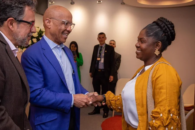 Francia Marquéz, Vicepresidenta de Colombia, una mujer afro latina con una chaqueta amarilla y una camisa blanca da un apretón de manos a Darren Walker, Presidente de la Fundación Ford, un hombre negro con un blazer azul y una camisa azul claro en la oficina de la Fundación Ford en Bogotá. A su derecha Javier Ciurlizza, Director Regional de la Fundación Ford, un hombre con lentes usando una chaqueta de traje marrón los mira y sonríe. Detrás de ellos hay paredes naranjas y blancas, flores y otras personas. 