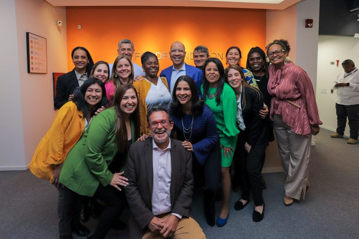 Un grupo de empleados de la Fundación Ford posan con Francia Marquéz, Vicepresidenta de de Colombia, una mujer afro latina con una chaqueta amarilla y una camisa blanca, y Darren Walker, Presidente de la Fundación Ford, un hombre negro con un blazer azul y una camisa azul claro, se abrazan en la oficina de la Fundación Ford en Bogotá. Detrás de ellos hay una pared naranja y el logo de la Fundación Ford. 