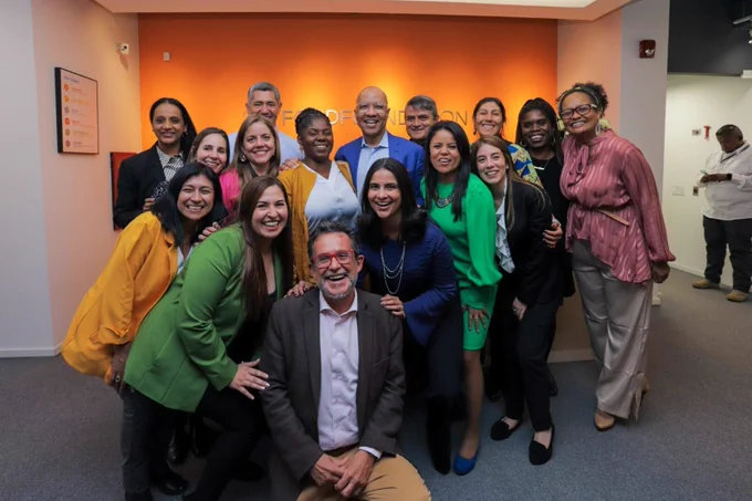 Un grupo de empleados de la Fundación Ford posan con Francia Marquéz, Vicepresidenta de de Colombia, una mujer afro latina con una chaqueta amarilla y una camisa blanca, y Darren Walker, Presidente de la Fundación Ford, un hombre negro con un blazer azul y una camisa azul claro, se abrazan en la oficina de la Fundación Ford en Bogotá. Detrás de ellos hay una pared naranja y el logo de la Fundación Ford. 