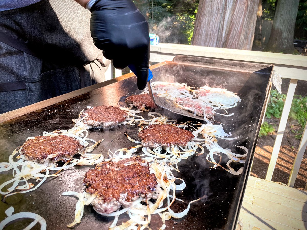 Smash burgers in the process of being flipped. There are onions on the griddle side of the patties and a serious crust on the top.