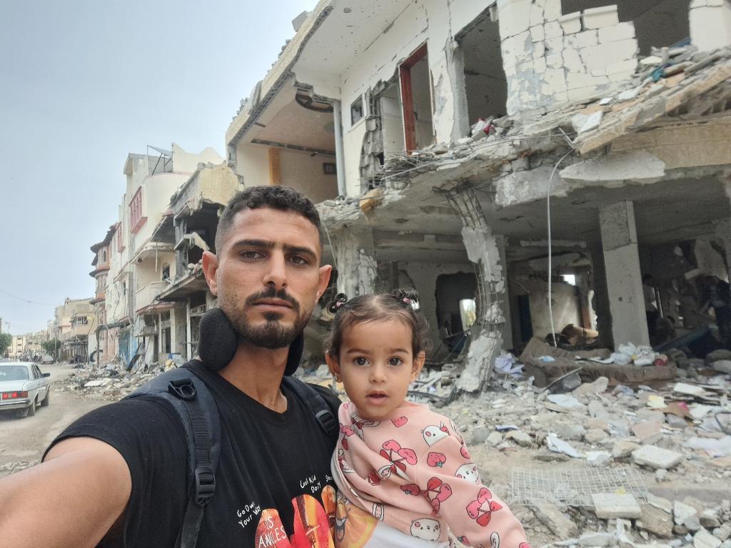 Me and my child in my destroyed town, Khuza’a. There were houses here, and here was everything. We will remain until we revive it. May God protect us. Your prayers for us.