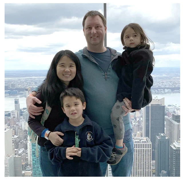 An Asian woman poses with a white man and two small children in front of a skyscraper window. New York City  is visible behind them. 