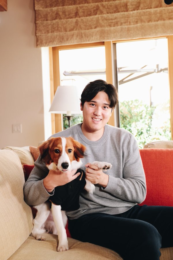 Shohei Ohtani smiles and sits on a couch with his dog next to him with the dog's paw in Ohtani's hand
