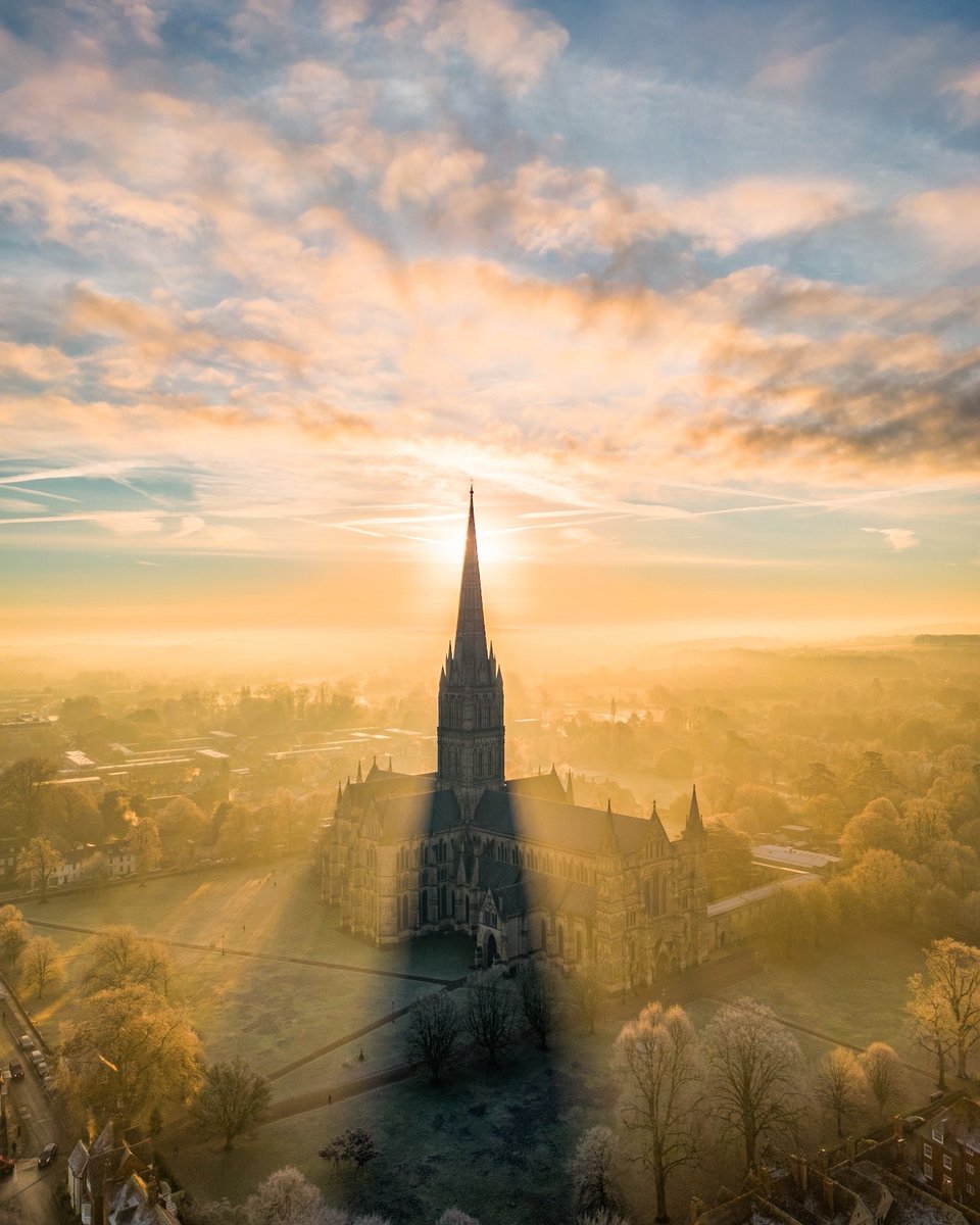 A drone aerial shot of Salisbury Cathedral as the sun is rising. The beams are casting over the surrounding green landscape and the Spire is creating a striking shadow.