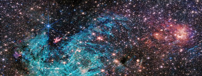 This image shows a field crowded with stars. A large, bright cyan-colored area surrounds the lower portion of a funnel-shaped region of space that is wider at the top edge of the image and then narrows. This funnel-shaped region appears darker than its surroundings. Toward the narrow end of this dark region a small clump of red and white appears to shoot out streamers upward and left. The cyan-colored area has needle-like structures and becomes more diffuse towards the right. The right side is dominated by clouds of orange and red, with a purple haze.