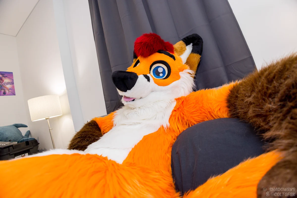 An orange fox fursuiter lying on a bed with his arms stretched out over the bed head.