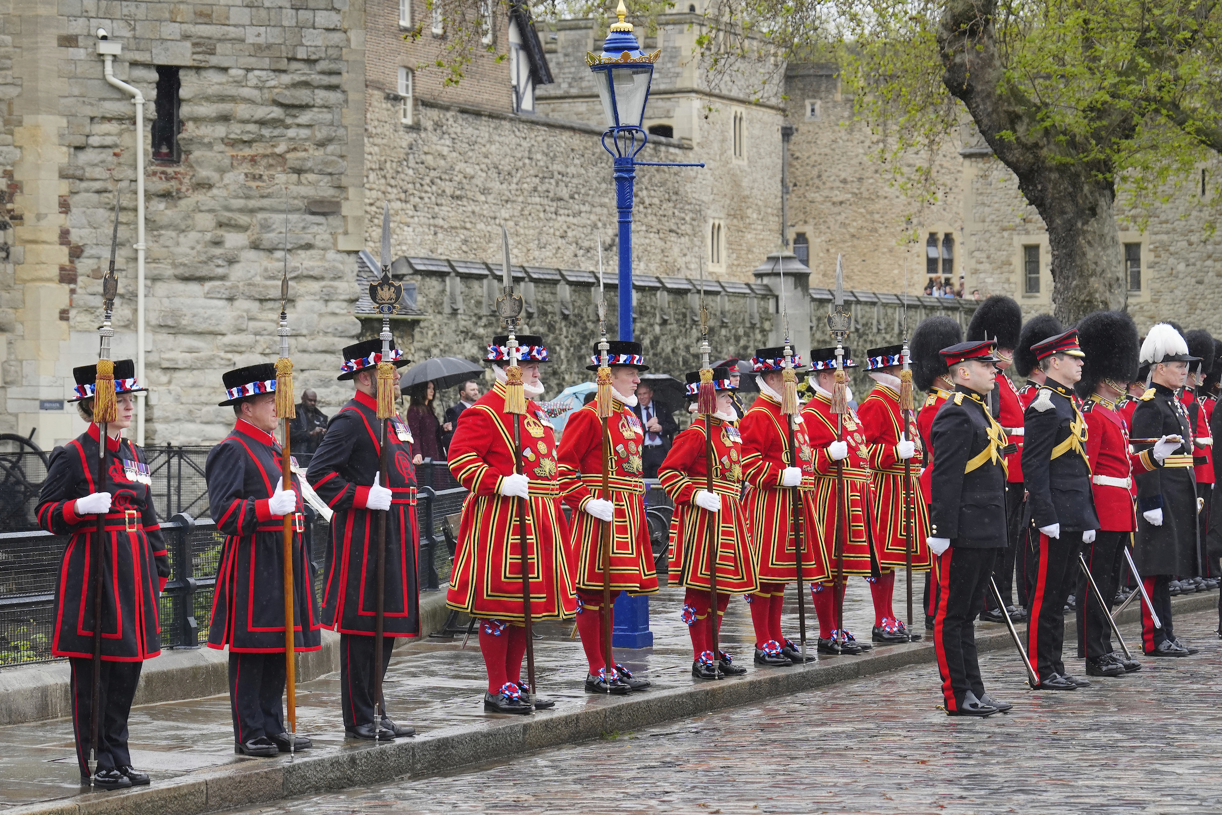 A row of Yeoman Warders wearing red state dress uniform and blue and red undress uniform. There is a stone building behind them. 