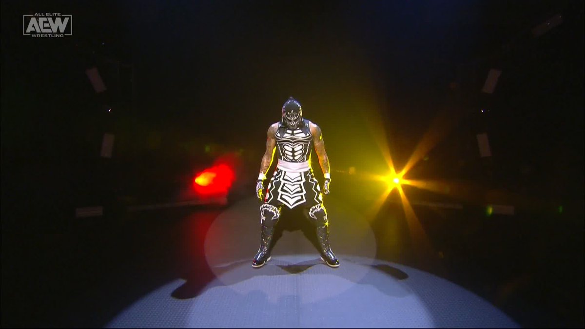 A wrestler in a black and white skeletal-themed mask and outfit stands under dramatic red and yellow stage spotlights in an arena, with the AEW (All Elite Wrestling) logo visible in the top-left corner.