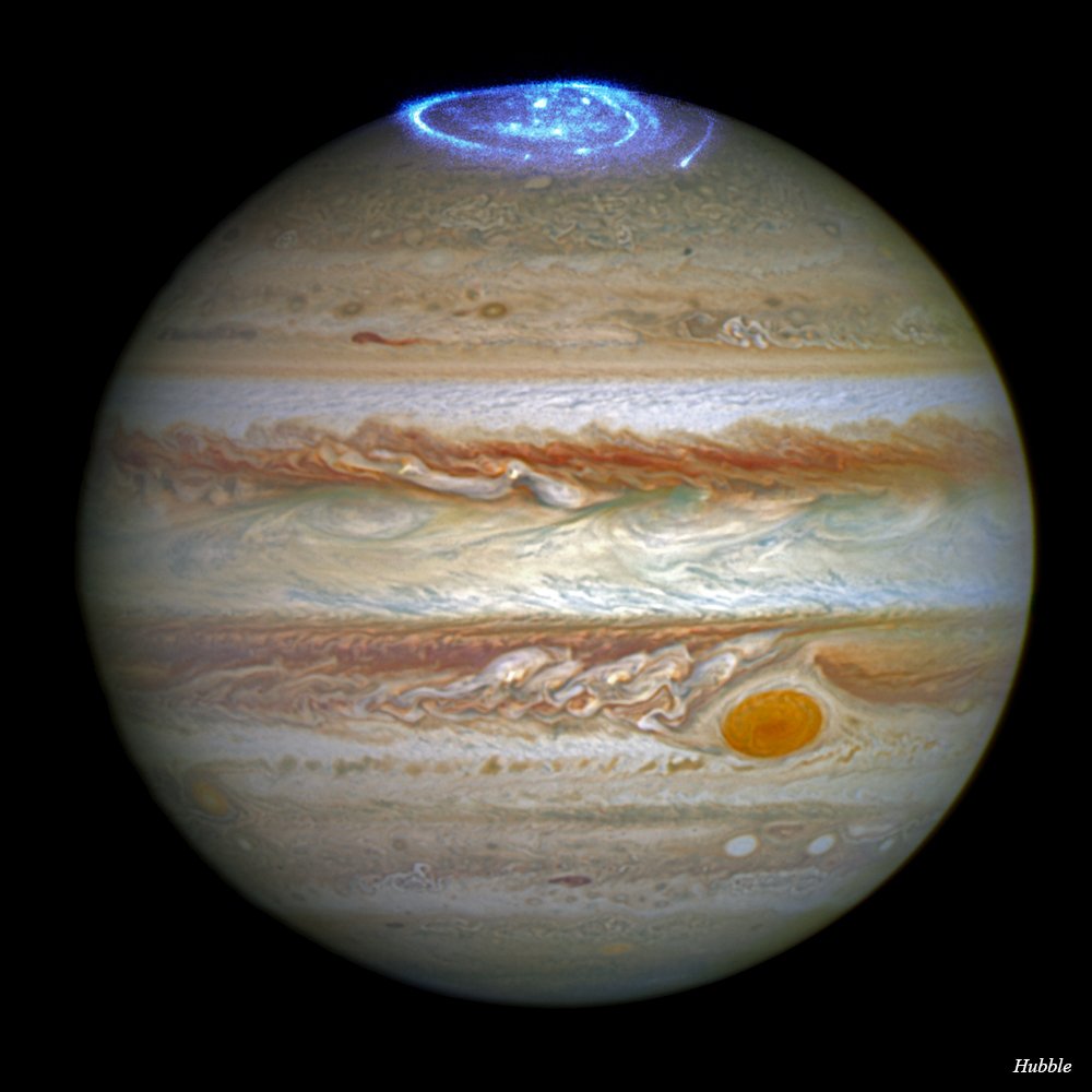 The planet Jupiter is seen here with stripes of orange, white, beige, and red. A halo of bright blue at the north pole are aurorae seen in ultraviolet light.