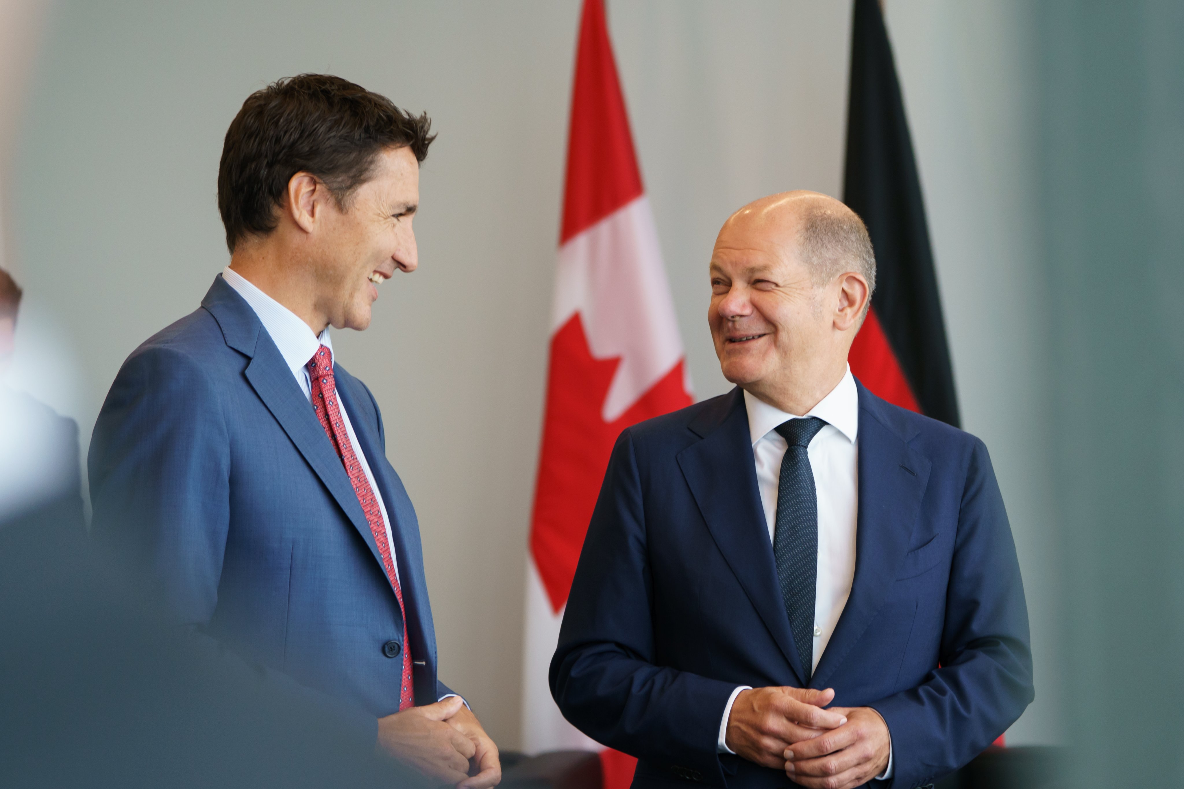 Prime Minister Justin Trudeau and German Chancellor Olaf Scholz are standing beside each other and smiling. The German and Canadian flags are visible behind them.