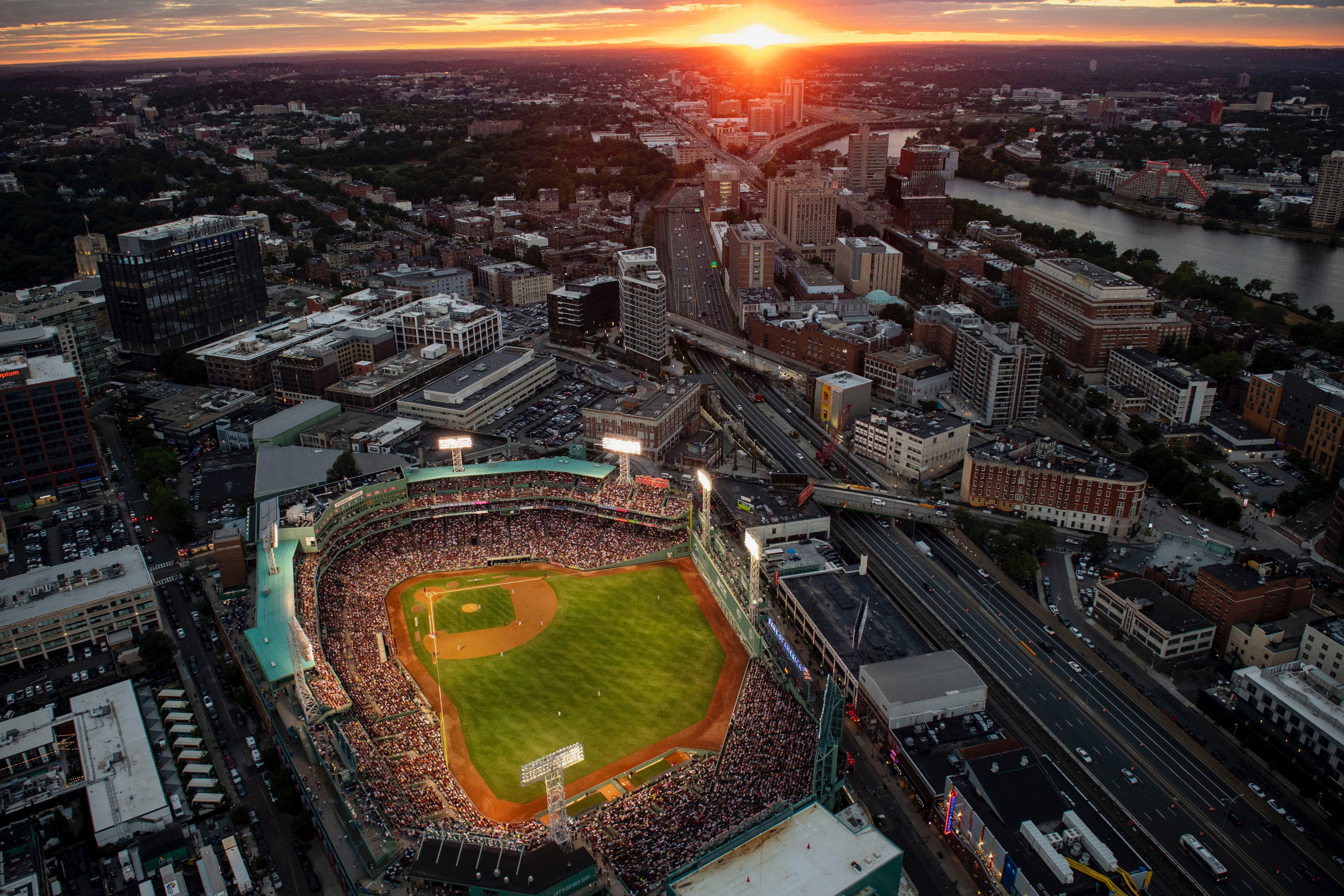 A wide shot of Fenway Park and the surrounding Boston neighborhoods and Charles River from a helicopter above the city at sunset. The sun sets on the horizon with the lights on at a sold-out Fenway Park during the game on 8/13/22. 