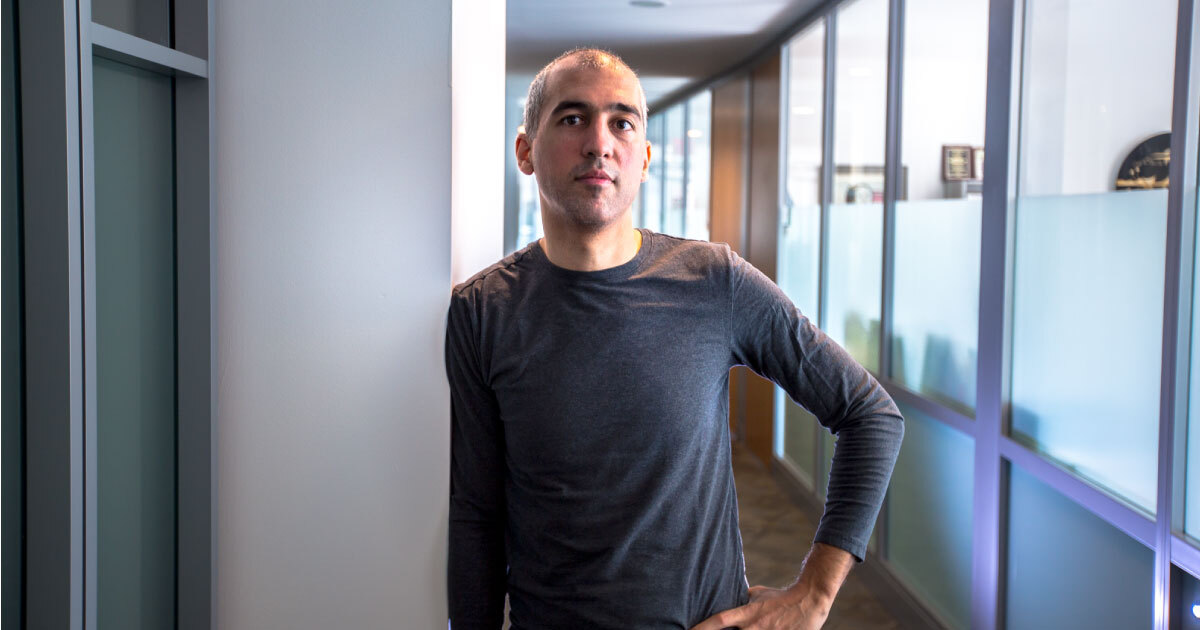 person standing in windowed hallway wearing a gray shirt with hand on hips posing for camera