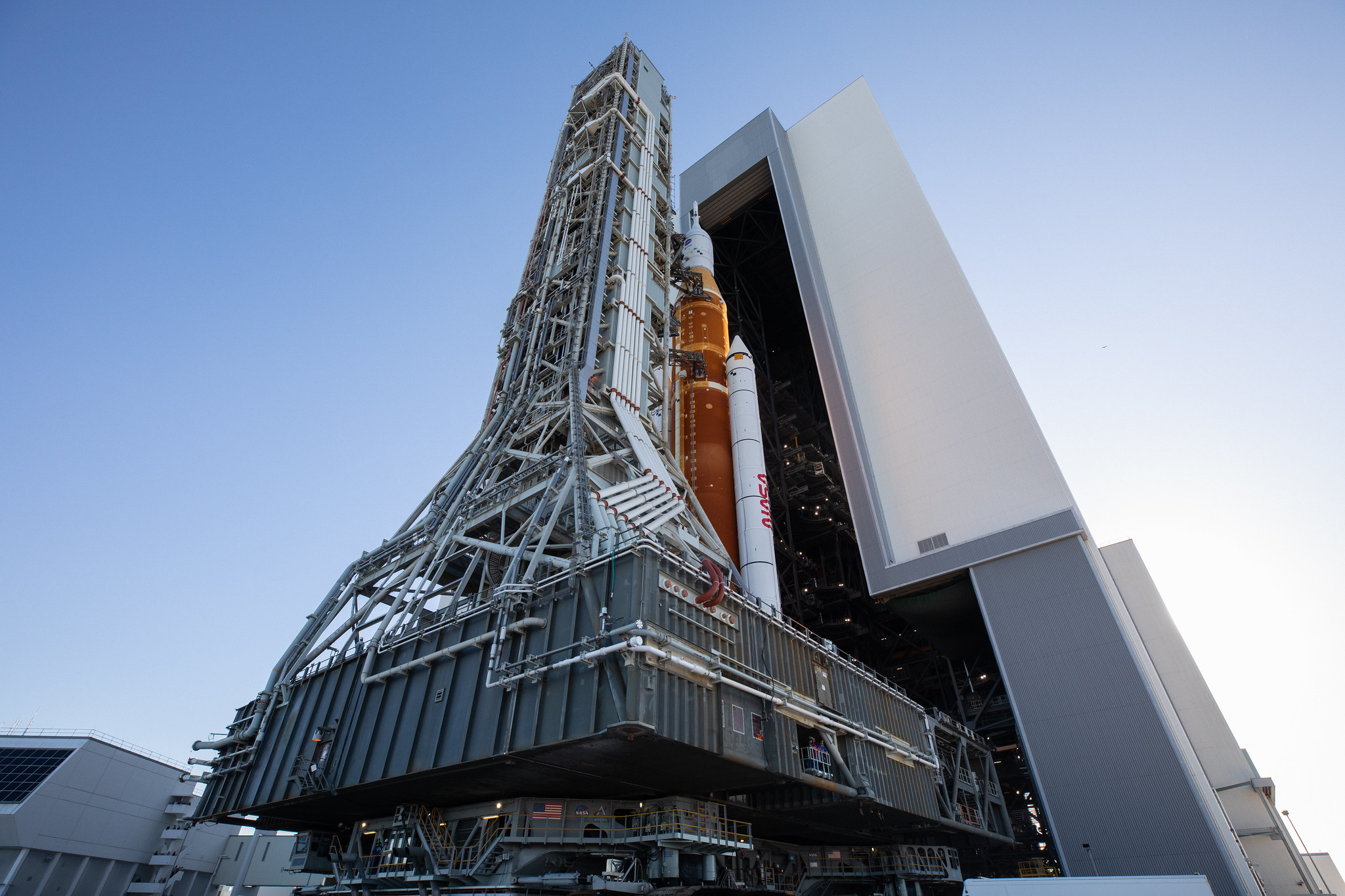 NASA's Space Launch System, a giant orange rocket with twin white boosters on each side and the white Orion spacecraft on top, rolls out of the Vehicle Assembly Building at NASA's Kennedy Space Center. This photo was taken from ground level looking sharply up at the rocket, which is standing on top of a large, square, dark gray transporter and is partially tucked behind a tall, light gray mobile launcher with countless tubes and wires.