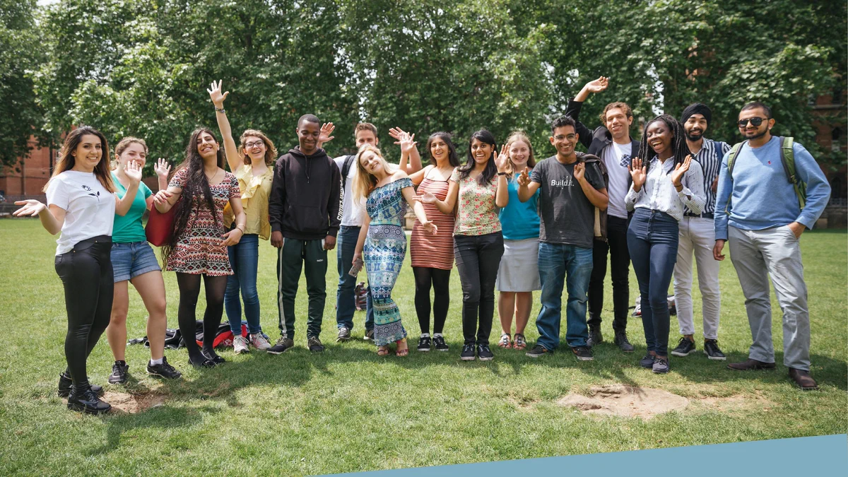 A group of students stand in a line in a grassy quad, throwing their arms in the air in celebration