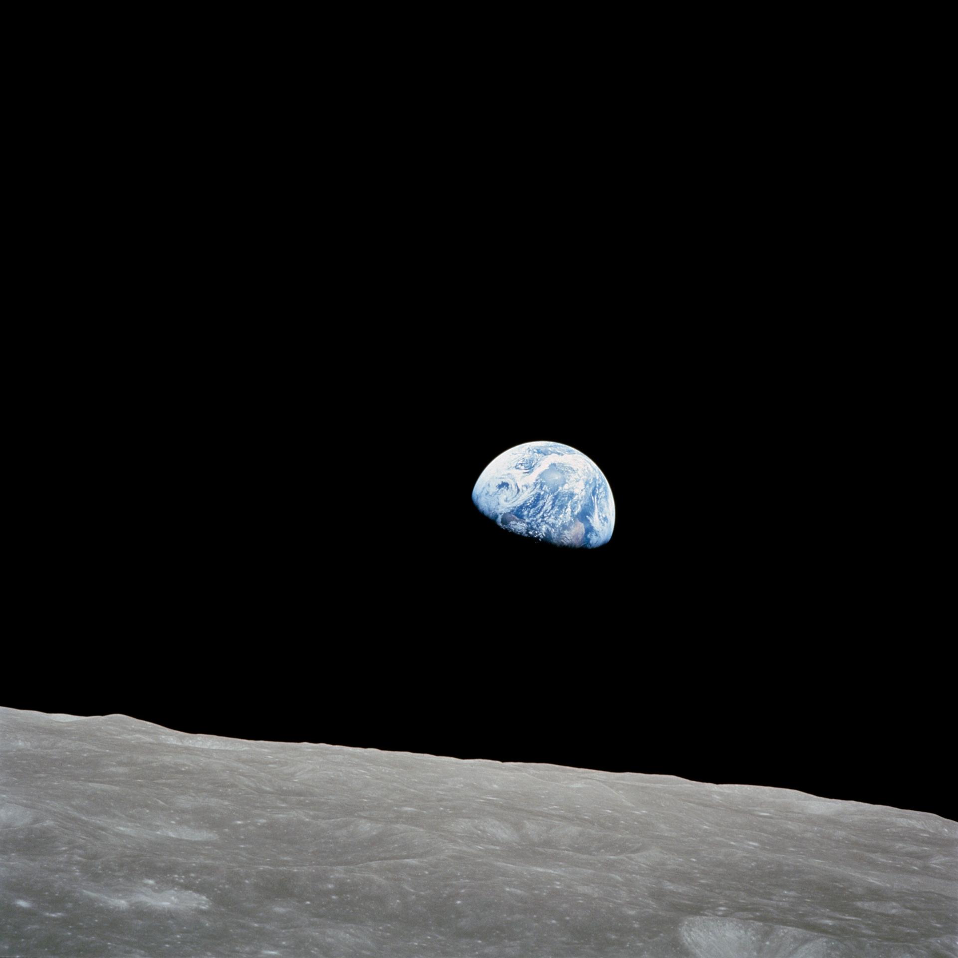 The rising Earth hangs about five degrees above a gray and barren lunar landscape. The planet's swirling white clouds and deep blue ocean waters stand out against the blackness of space.