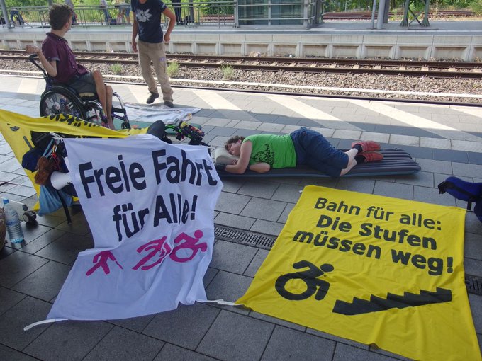 Menschen am Bahnsteig. Eine person im Rollstuhl, eine andere liegt auf Isomatte. Nanner freie Fahrt fürcalkes, bahn für alle die Stufen müssen weg