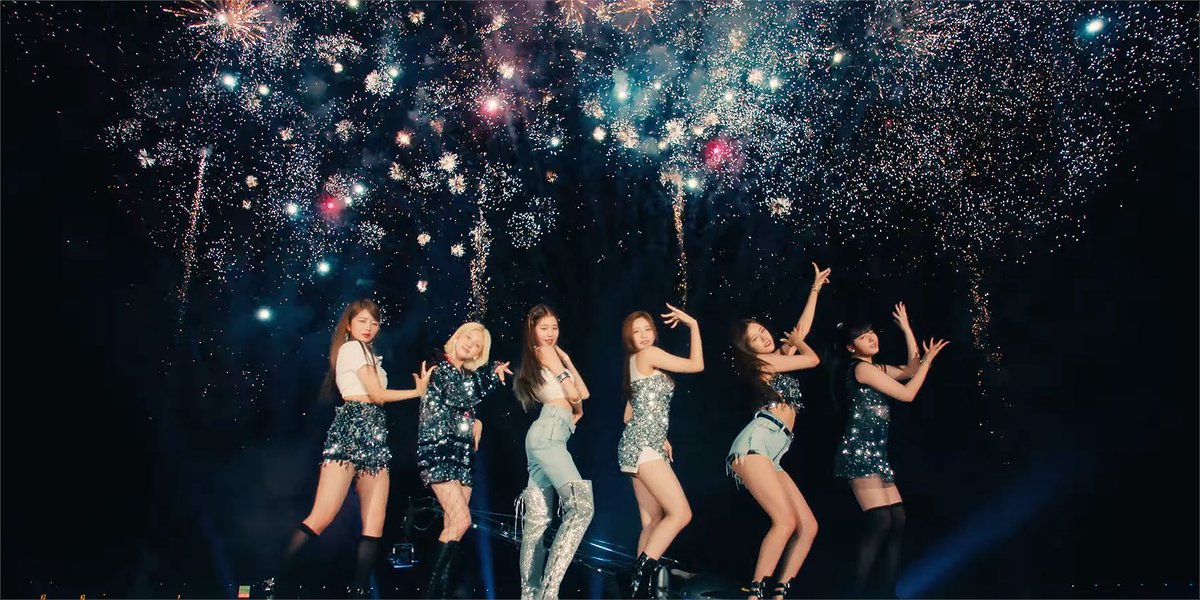 Six female performers in sparkly outfits pose on stage beneath an elaborate fireworks display lighting up the night sky.