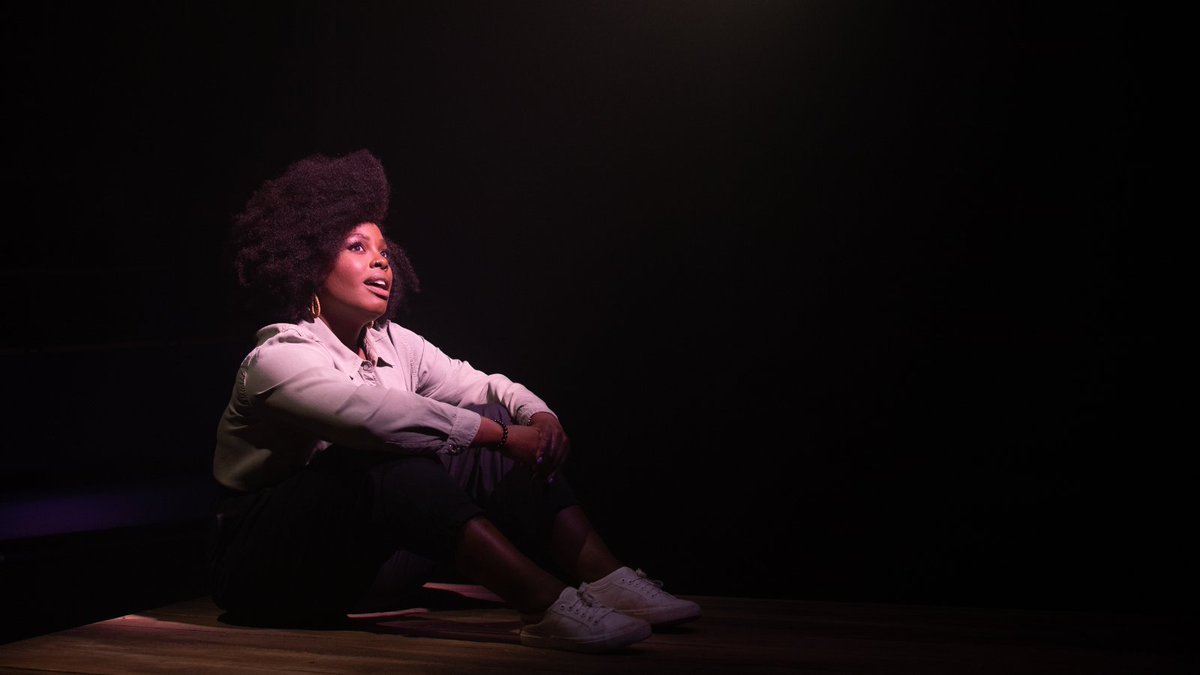 Nadine Higgin sits, wearing a shirt, black trousers and white trainers sits on a wooden floor, looking up towards the light. 