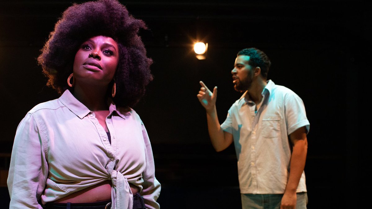 Nadine Higgin stands, wearing a shirt tied at the centre. She looks up and outwards. In the background, Aaron Anthony speaks out with his hand pointing in front of him. 