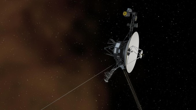 In this illustration, the Voyager 1 spacecraft appears on the right against a black background punctuated by stars. The spacecraft's large, white radio antenna faces to the right. Four booms extend from the spacecraft, three toward the bottom and one toward the top. On the left side of the view is a brown haze that partially obscures the background stars.