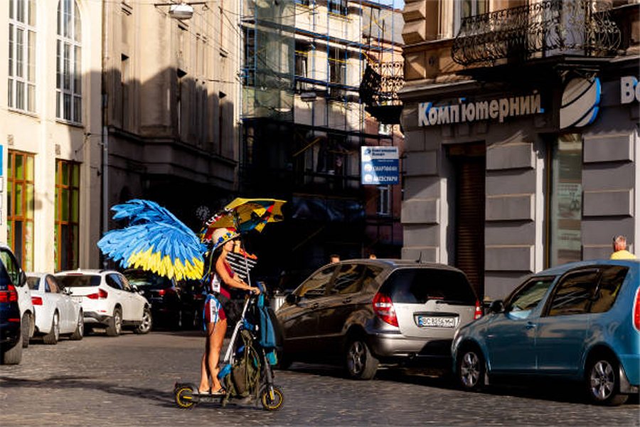zav_raya's tweet photo. A woman dressed with blue and yellow (Ukrainian national colours) wings rides a scooter in the old town of Lviv. #Ukraine️ #war #RussianAggression #RussianWarCrimes #RussiaIsATerroristState https://t.co/BKBs1qHheo