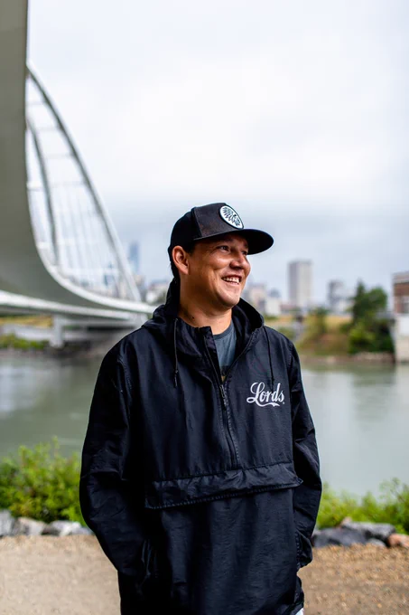 A deadly uncle standing by the river with a hat on looking off to their left smiling, with a bridge and city skyline across the river in the background.