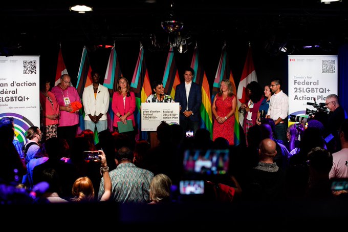 Minister Marci Ien is standing at a podium and speaking. Prime Minister Justin Trudeau, Minister Mona Fortier, Members of Parliament Jenna Sudds, Anita Vandenbeld, Marie-France LaLonde, and Yasir Naqvi, and community activists are also standing on stage. Behind them, Canada flags and Pride flags are visible.