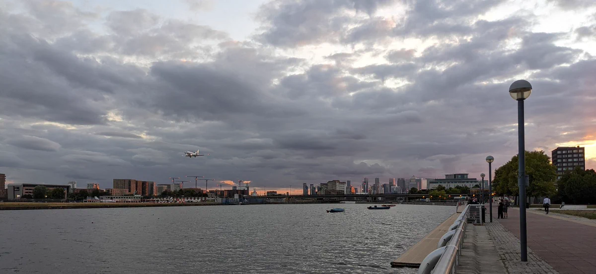 A view from Newham Dockside looking towards the City of London. The buildings in the City are lit on the north side by a low orange evening sun. A small private jet can be seen low on the left hand side coming in to land at London City Airport.
