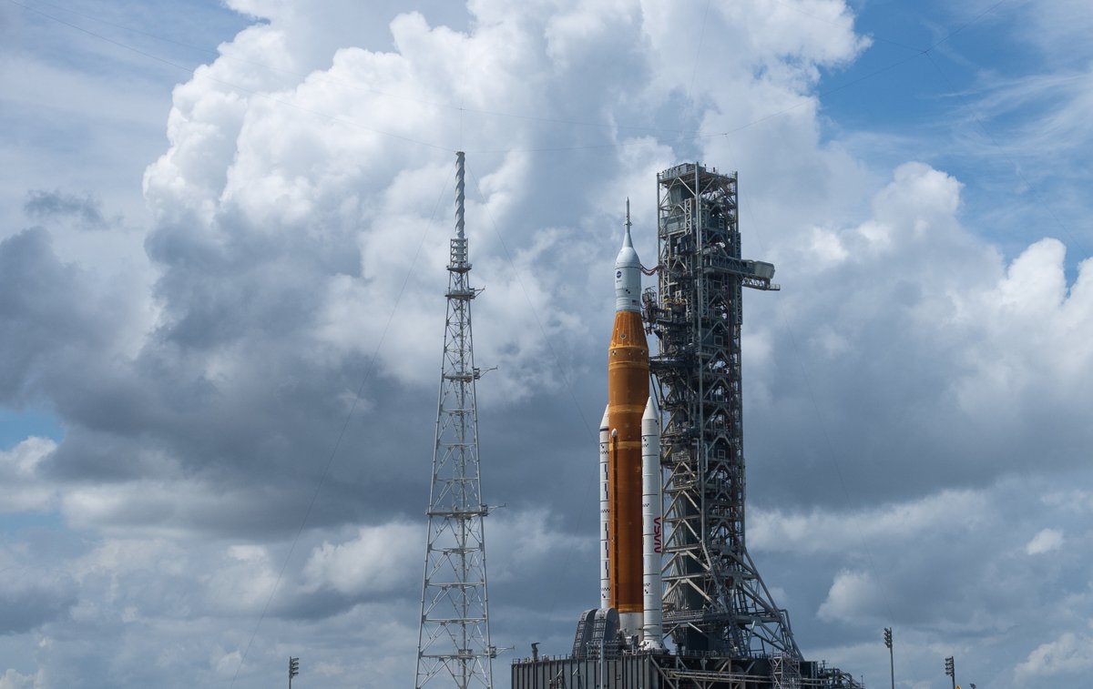 The Space Launch System rocket stands upright on the launchpad. The background is the sky dominated by puffy cumulus clouds. The rocket has an orange central fuel tank with two white rocket boosters on either side. The boosters have the word NASA painted vertically on them in red in the iconic "NASA Worm" typeface. The typeface resembles a worm. Photo credit: NASA/Joel Kowsky